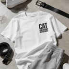A white short-sleeve T-shirt laid flat on a light stone surface, printed with the words 'CAT EMERGENCY DEPARTMENT' in black text on the chest. Gray sweatpants, a black belt, and white sneakers are styled around it as outfit props.