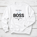 A white crewneck sweatshirt with a graphic print that reads 'THE REAL BOSS lives here.' in black text, featuring a small pastel cat outline and paw print, displayed flat on a white wood plank background.