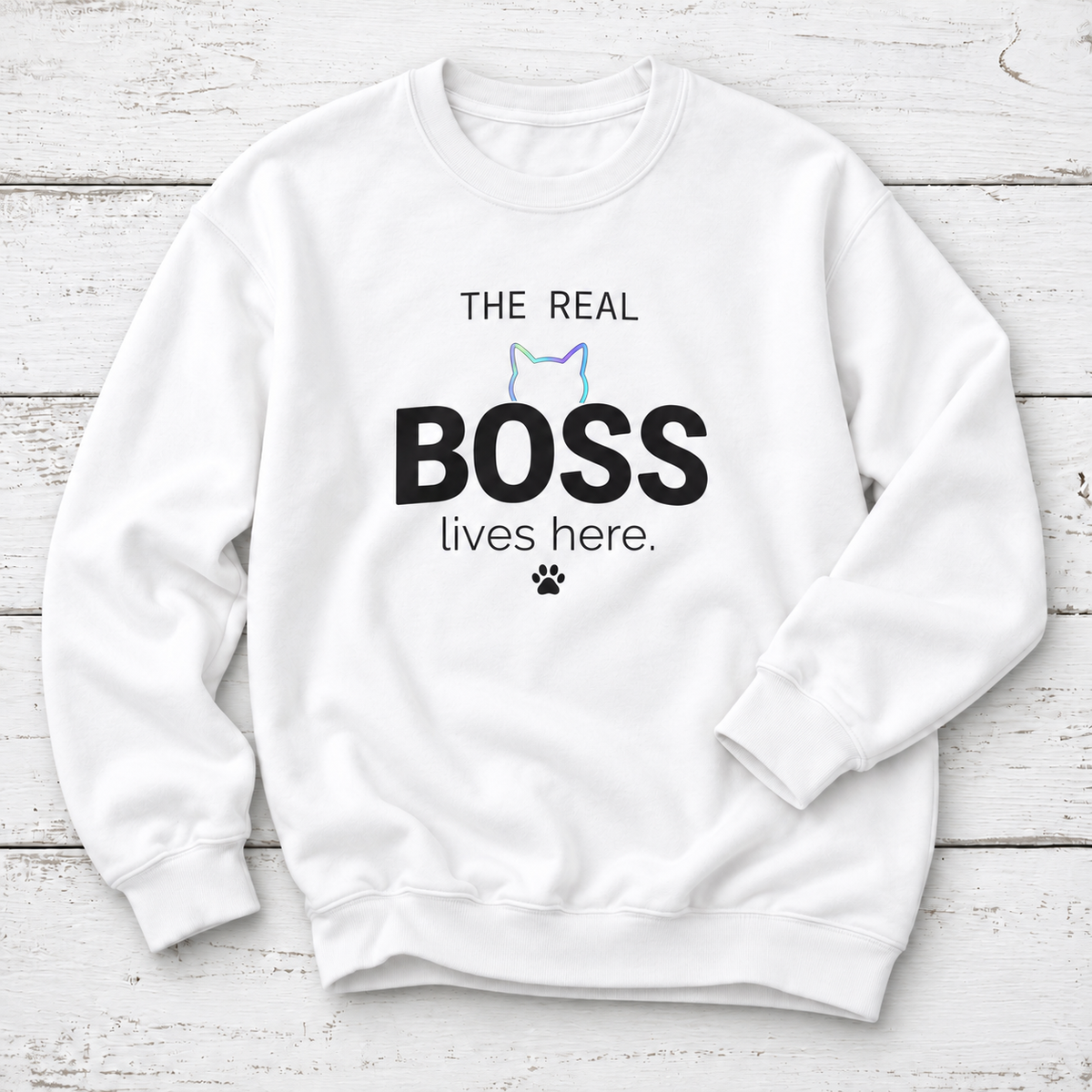 A white crewneck sweatshirt with a graphic print that reads 'THE REAL BOSS lives here.' in black text, featuring a small pastel cat outline and paw print, displayed flat on a white wood plank background.