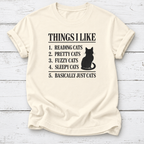 Cream T-shirt with a black cat silhouette and “Things I Like” list ending in “Basically Just Cats,” laid flat on white wooden boards.