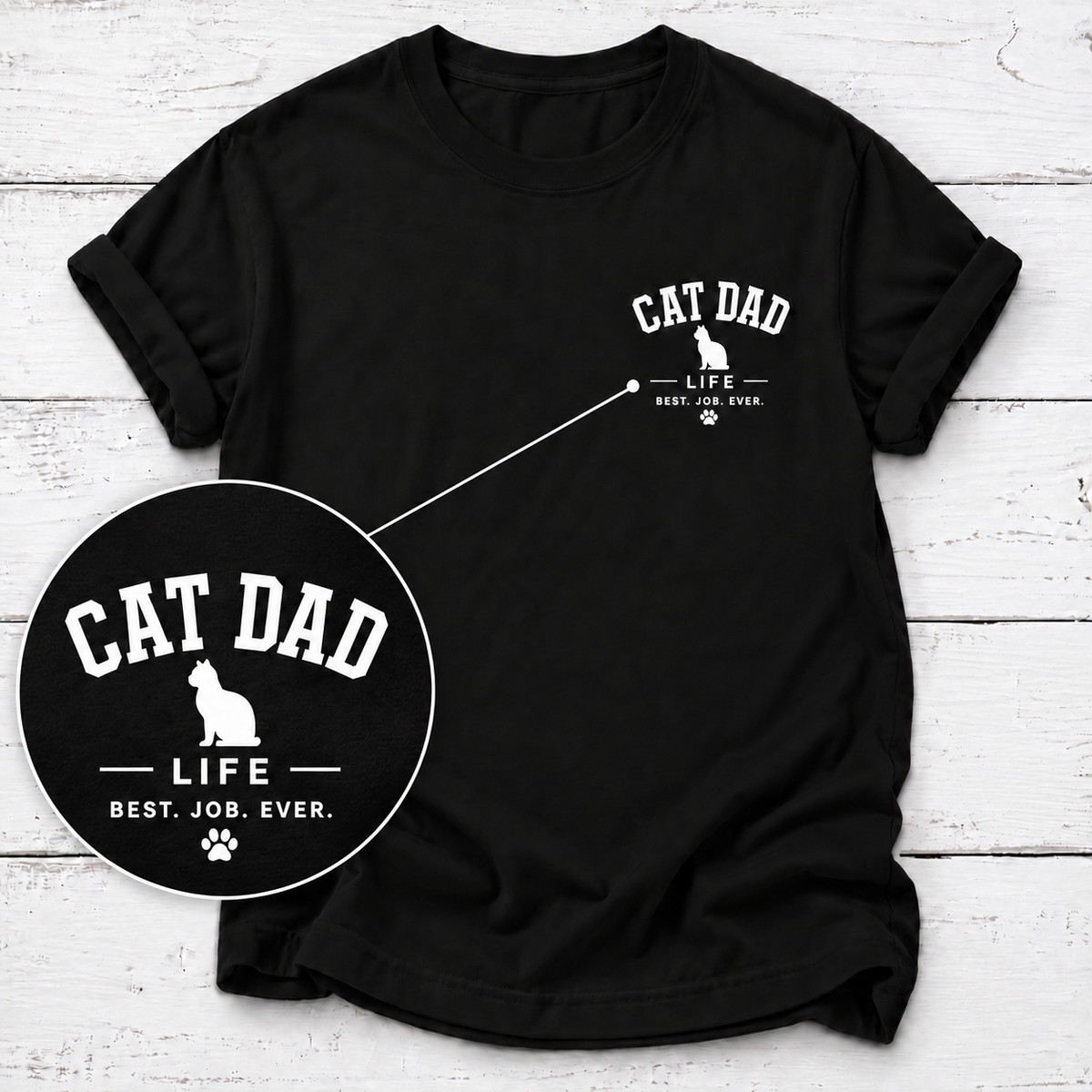 A black short-sleeve t-shirt with a white 'CAT DAD' graphic on the left chest, displayed on a white wood plank background with a circular zoom-in detail overlay.
