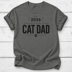 A dark gray crewneck t-shirt with a black graphic print that reads 'EST. 2026 OFFICIAL CAT DAD' with a paw print, laid flat on a white wood plank background.