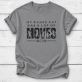 A gray short-sleeve crewneck t-shirt with a black graphic print that reads 'MY DANCE CAT HAS A LOT OF MOVES' with cat silhouettes inside the letters 'M-O-V-E-S' and a heartbeat line. The t-shirt is laid flat on a white wood plank background.