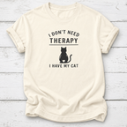 An off-white short-sleeve t-shirt with a black graphic print that reads 'I DON'T NEED THERAPY' above a black cat silhouette and 'I HAVE MY CAT' below, displayed flat on a white wood plank background.