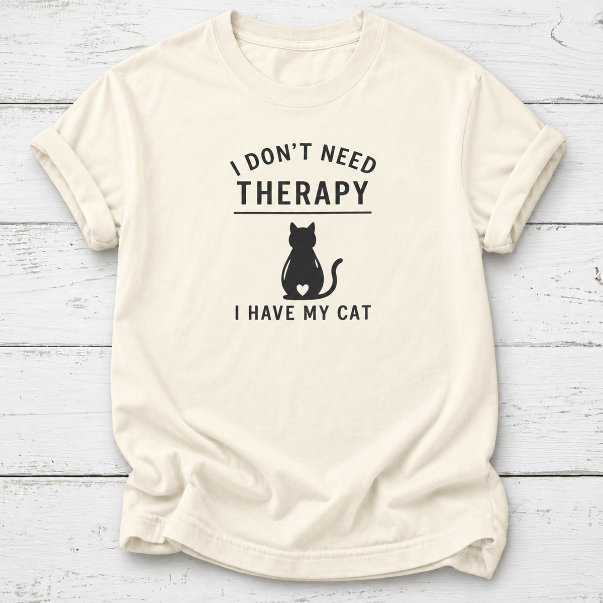 An off-white short-sleeve t-shirt with a black graphic print that reads 'I DON'T NEED THERAPY' above a black cat silhouette and 'I HAVE MY CAT' below, displayed flat on a white wood plank background.