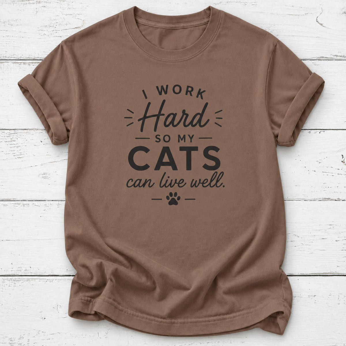 A brown crewneck t-shirt with a black graphic print that reads 'EST. 2026 OFFICIAL CAT DAD' with a paw print, laid flat on a white wood plank background.