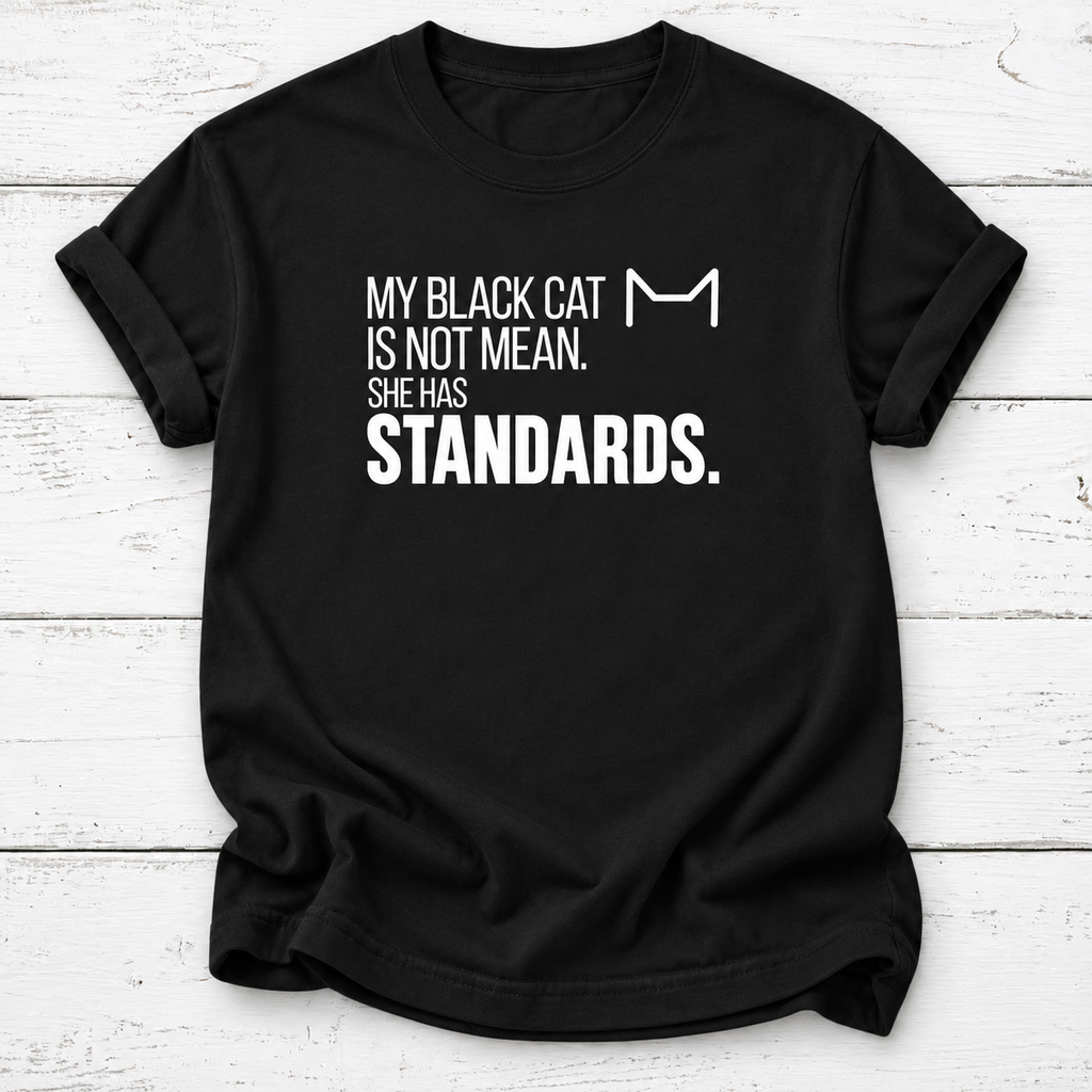 Black T-shirt with white “My Black Cat Is Not Mean. She Has Standards.” lettering and a minimalist cat-ear outline, laid flat on white wooden boards.