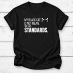 Black T-shirt with white “My Black Cat Is Not Mean. She Has Standards.” lettering and a minimalist cat-ear outline, laid flat on white wooden boards.