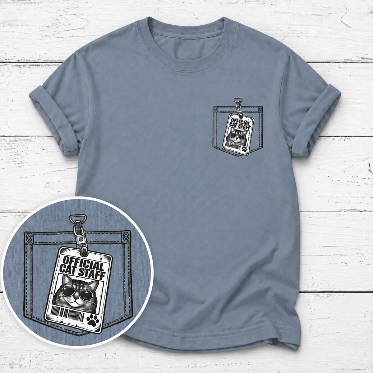 A dusty slate blue short-sleeve t-shirt with a graphic print of a cat ID badge styled as a pocket label on the left chest. A circular inset shows a close-up of the graphic design, which reads 'OFFICIAL CAT STAFF' with a grumpy cat face and paw print.