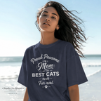 A woman wearing an oversized navy graphic T-shirt on a beach. The shirt features white text that reads 'Proud Pawsome Mom of the BEST CATS ever Fur real.