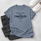 A light blue graphic t-shirt with the text 'COFFEE, CATS, AND CANCELED PLANS' and a coffee cup icon, styled in a flat lay with black jeans and white sneakers on a light gray background.