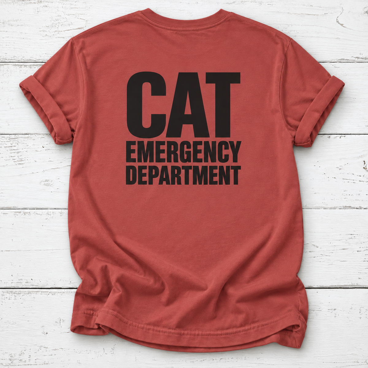 A rust-red t-shirt with 'CAT EMERGENCY DEPARTMENT' printed in bold black letters on the back, laid flat on a white wood plank background.