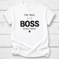 A white short-sleeve crewneck t-shirt laid flat on a white wood plank background. The shirt features a graphic print that reads 'THE REAL BOSS lives here.