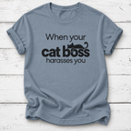 Dusty blue T-shirt with “When your cat boss harasses you” lettering and a lounging black cat graphic, laid flat on white wooden boards