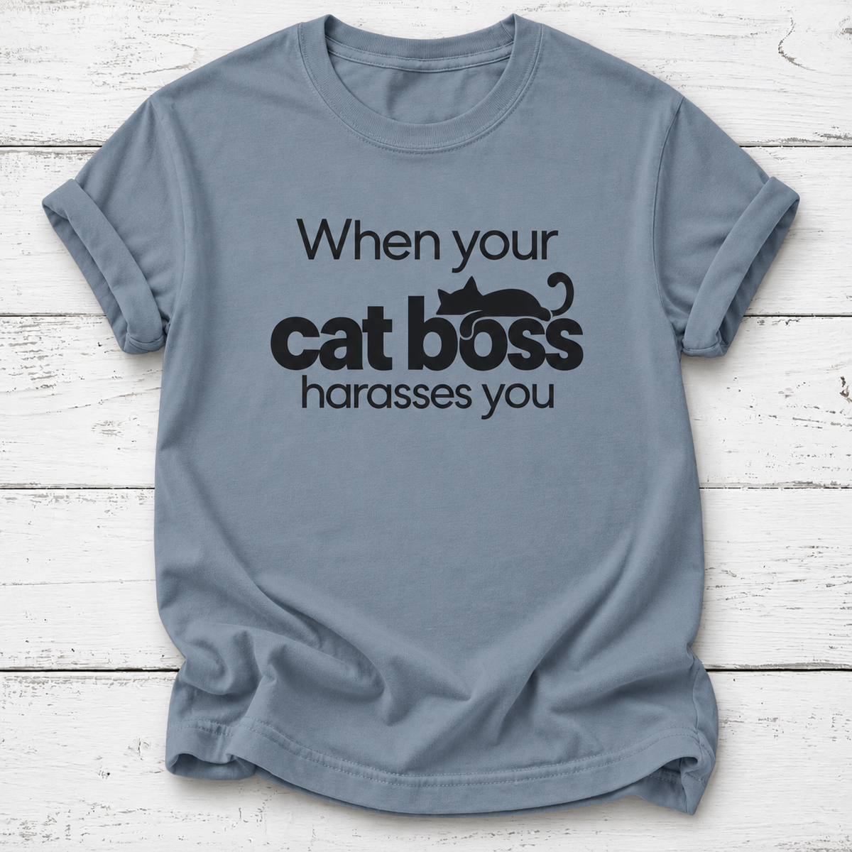 Dusty blue T-shirt with “When your cat boss harasses you” lettering and a lounging black cat graphic, laid flat on white wooden boards