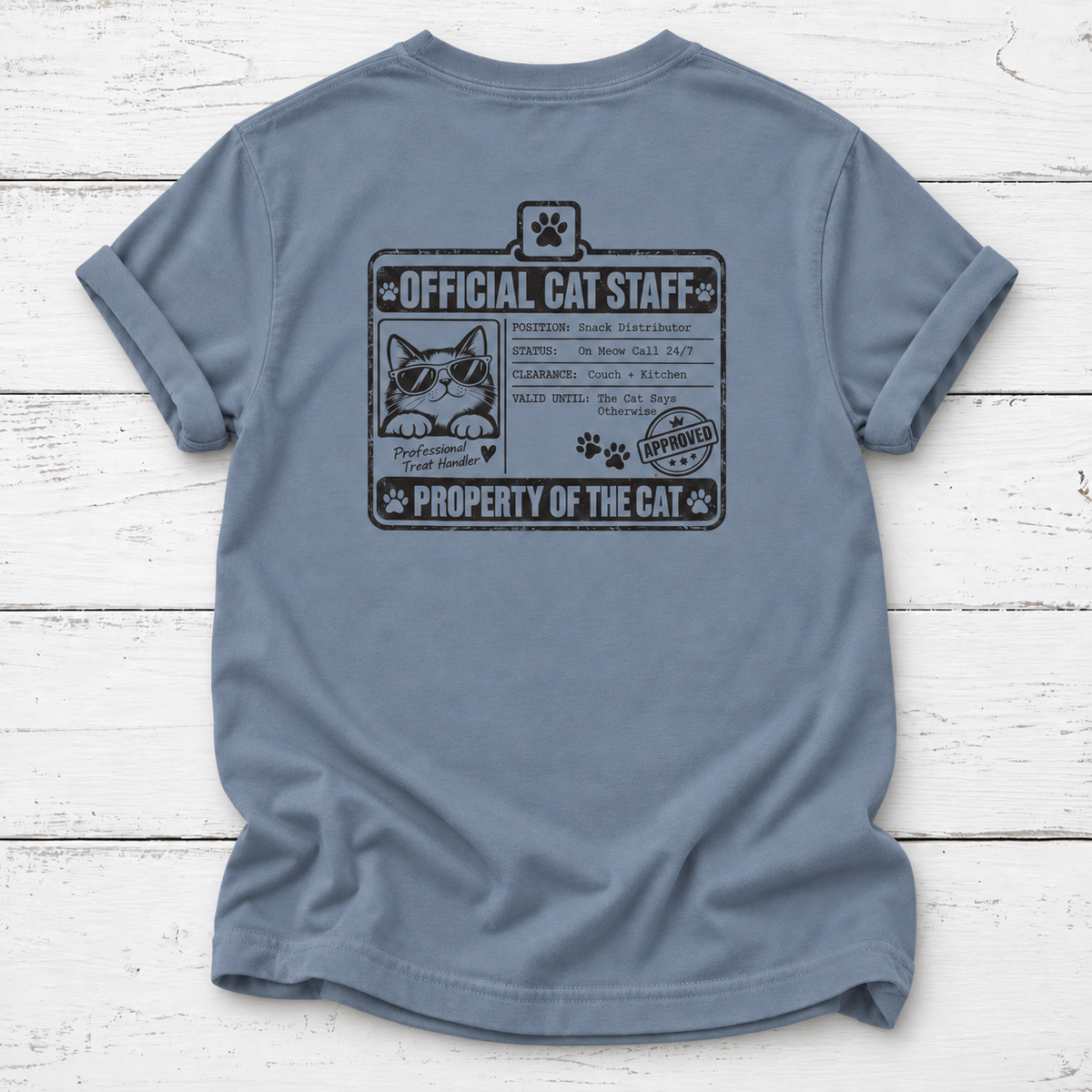 A slate blue short-sleeve t-shirt laid flat on a white wood plank background, featuring a black graphic print of an 'Official Cat Staff' badge on the front.