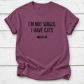 A mauve-purple short-sleeve t-shirt with black text reading 'I'M NOT SINGLE, I HAVE CATS' and a fish skeleton graphic, laid flat on a white wood plank background.