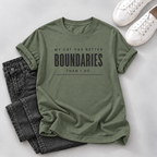 An olive green graphic t-shirt with the text 'MY CAT HAS BETTER BOUNDARIES THAN I DO.' printed in black, styled in a flat lay with dark gray jeans and white sneakers on a light gray background.