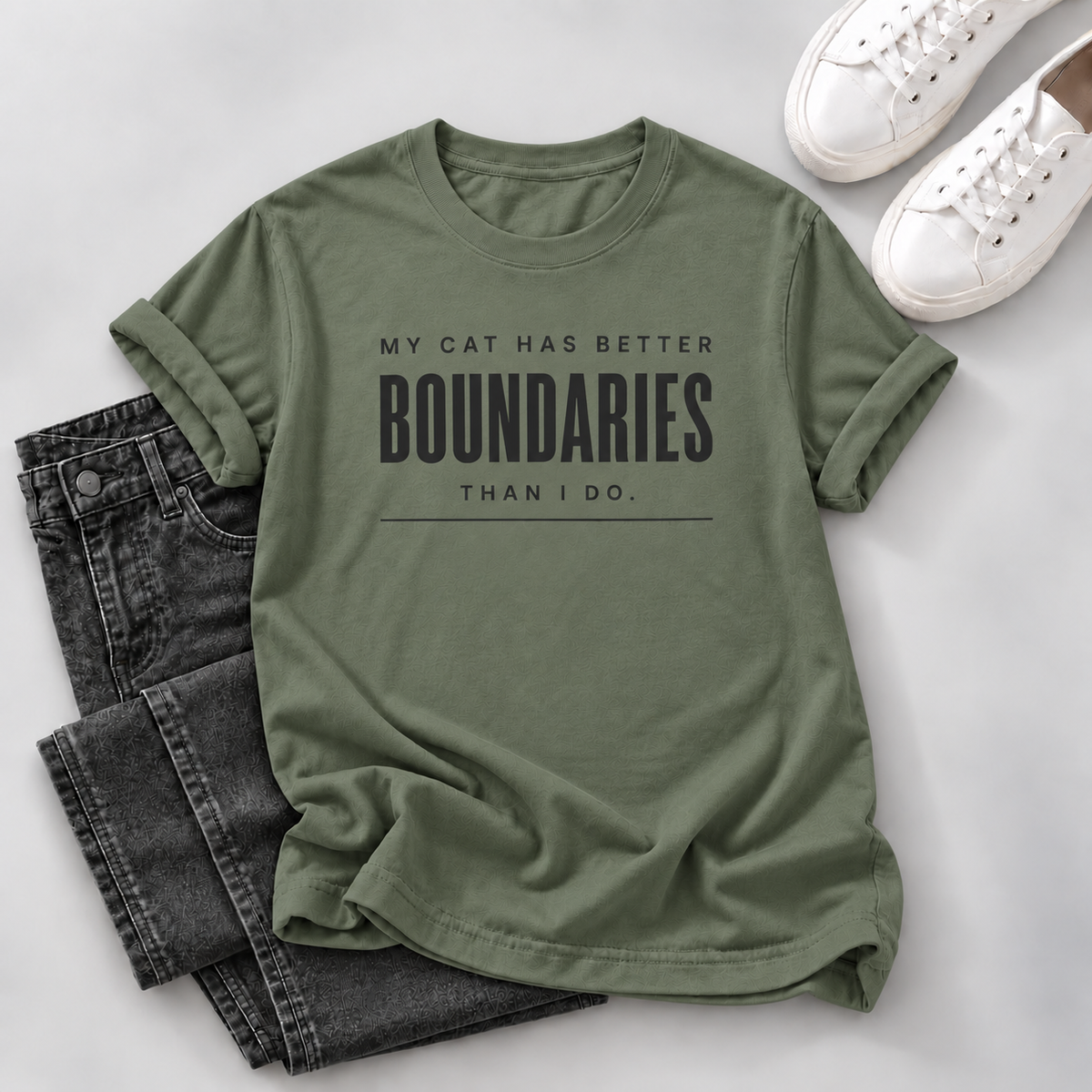 An olive green graphic t-shirt with the text 'MY CAT HAS BETTER BOUNDARIES THAN I DO.' printed in black, styled in a flat lay with dark gray jeans and white sneakers on a light gray background.