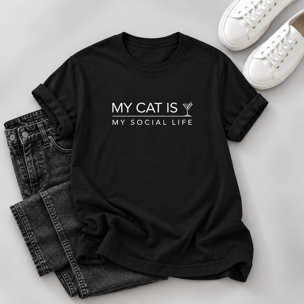 A black short-sleeve graphic T-shirt laid flat on a light gray surface, styled with folded dark jeans and white sneakers. The shirt has white text reading 'MY CAT IS MY SOCIAL LIFE' with a small cocktail glass icon.