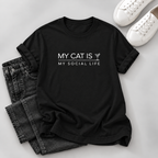 A black short-sleeve graphic T-shirt laid flat on a light gray surface, styled with folded dark jeans and white sneakers. The shirt has white text reading 'MY CAT IS MY SOCIAL LIFE' with a small cocktail glass icon.
