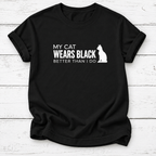 A black short-sleeve t-shirt with a white graphic print that reads 'MY CAT WEARS BLACK BETTER THAN I DO' alongside a white cat silhouette, displayed on a white wood plank background.