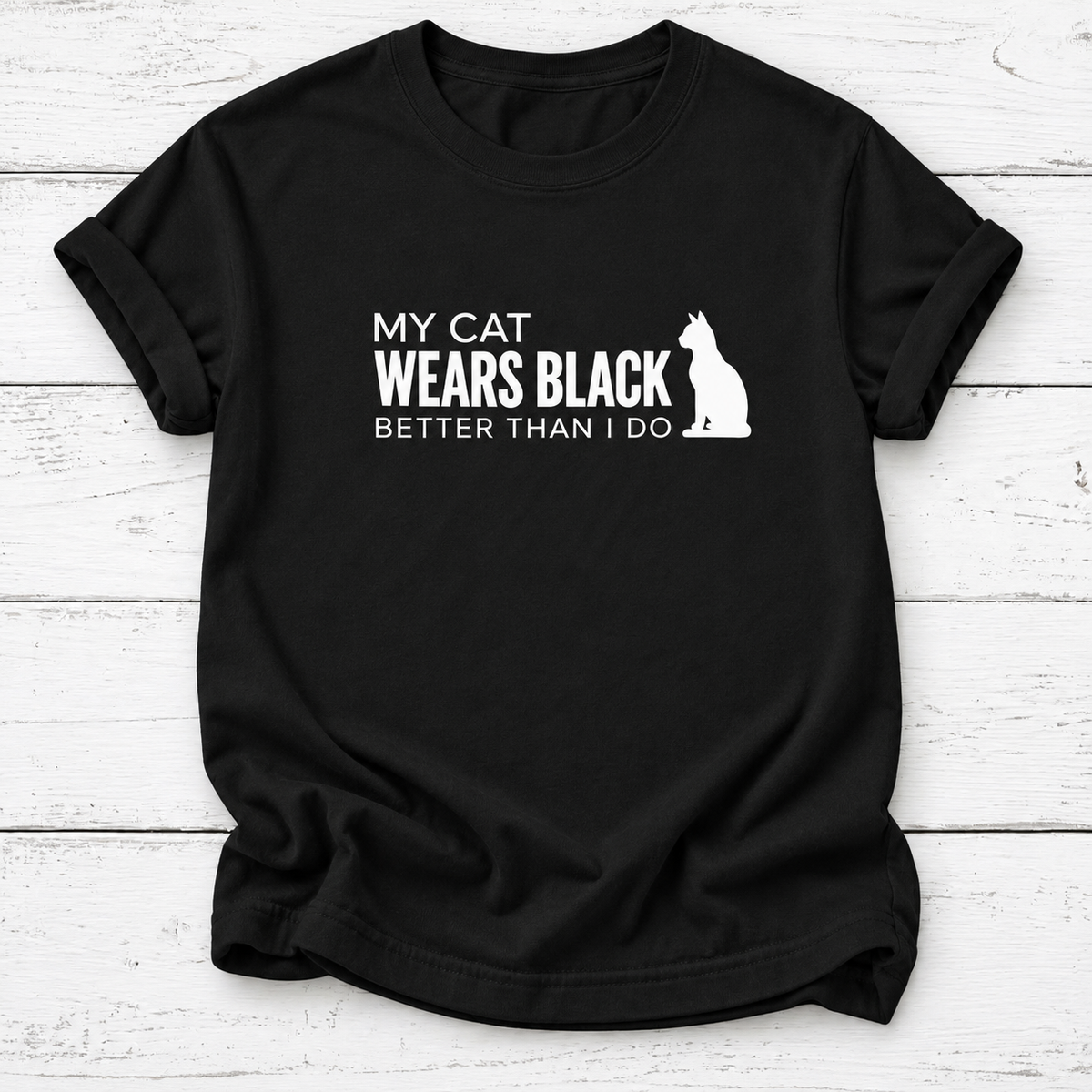 A black short-sleeve t-shirt with a white graphic print that reads 'MY CAT WEARS BLACK BETTER THAN I DO' alongside a white cat silhouette, displayed on a white wood plank background.