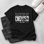A black short-sleeve graphic T-shirt laid flat on a light gray surface, styled with folded dark gray jeans and white sneakers. The shirt has a white printed slogan reading “MY DANCE CAT HAS A LOT OF MOVES” with cat silhouettes and an EKG line graphic.