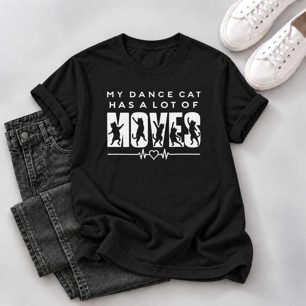 A black short-sleeve graphic T-shirt laid flat on a light gray surface, styled with folded dark gray jeans and white sneakers. The shirt has a white printed slogan reading “MY DANCE CAT HAS A LOT OF MOVES” with cat silhouettes and an EKG line graphic.
