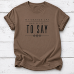 A brown short-sleeve crewneck t-shirt with the text 'MY ORANGE CAT HAS A LOT TO SAY' printed in black on the front, laid flat on a white wood plank background.
