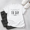 A white short-sleeve graphic T-shirt laid flat on a light gray surface, styled with folded dark gray jeans and white sneakers. The shirt has black text reading 'MY ORANGE CAT HAS A LOT TO SAY' with a soundwave graphic below.