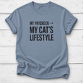 A dusty blue crewneck t-shirt with the text 'MY PAYCHECK -> MY CAT'S LIFESTYLE' printed in black on the front, laid flat on a white wood plank background.