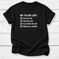 A black short-sleeve t-shirt with a white printed graphic that reads 'MY TO-DO LIST:' followed by four checked items: 'PET MY CAT', 'FEED MY CAT', 'PLAY WITH MY CAT', and 'REPEAT ALL ABOVE.' The shirt is laid flat on a white wood-plank background.