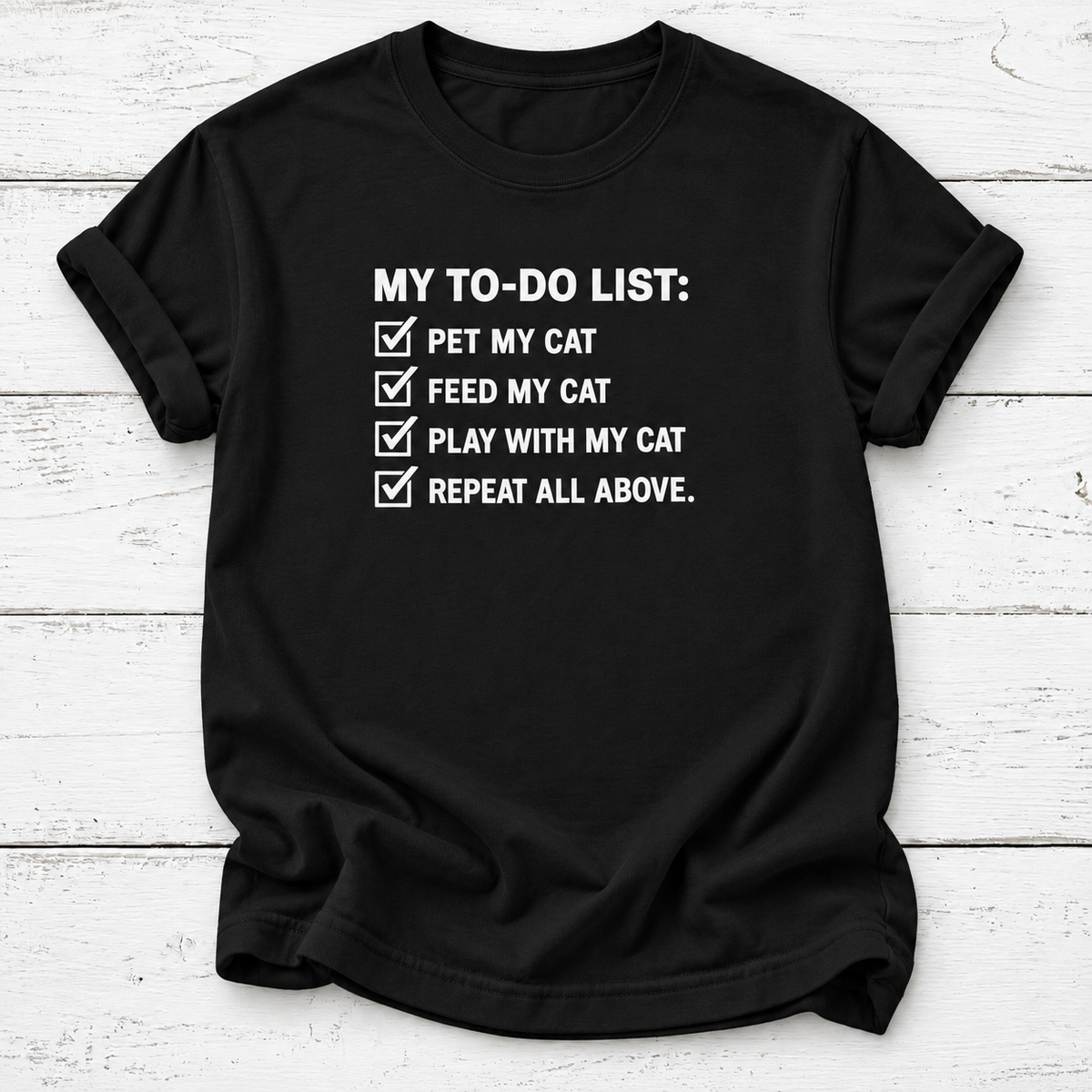 A black short-sleeve t-shirt with a white printed graphic that reads 'MY TO-DO LIST:' followed by four checked items: 'PET MY CAT', 'FEED MY CAT', 'PLAY WITH MY CAT', and 'REPEAT ALL ABOVE.' The shirt is laid flat on a white wood-plank background.