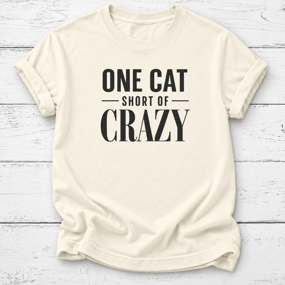 An off-white short-sleeve t-shirt with rolled sleeves, featuring the text 'ONE CAT SHORT OF CRAZY' printed in black on the front, laid flat on a white wood plank background.