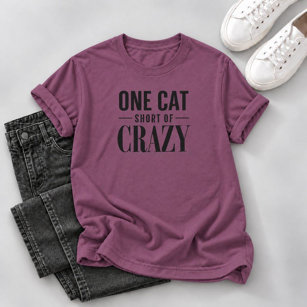 A mauve-purple short-sleeve t-shirt with black text reading 'ONE CAT SHORT OF CRAZY' is styled in a flat lay with dark gray jeans and white sneakers on a light gray background.