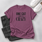 A mauve-purple short-sleeve t-shirt with black text reading 'ONE CAT SHORT OF CRAZY' is styled in a flat lay with dark gray jeans and white sneakers on a light gray background.